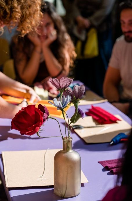 Un florero con flores de colores sobre una mesa con personas al fondo.